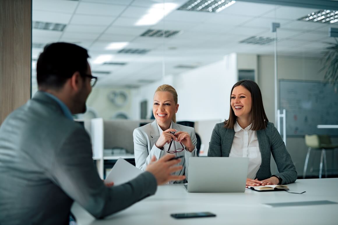 two smiling women and a man sitting at a desk with a laptop open 