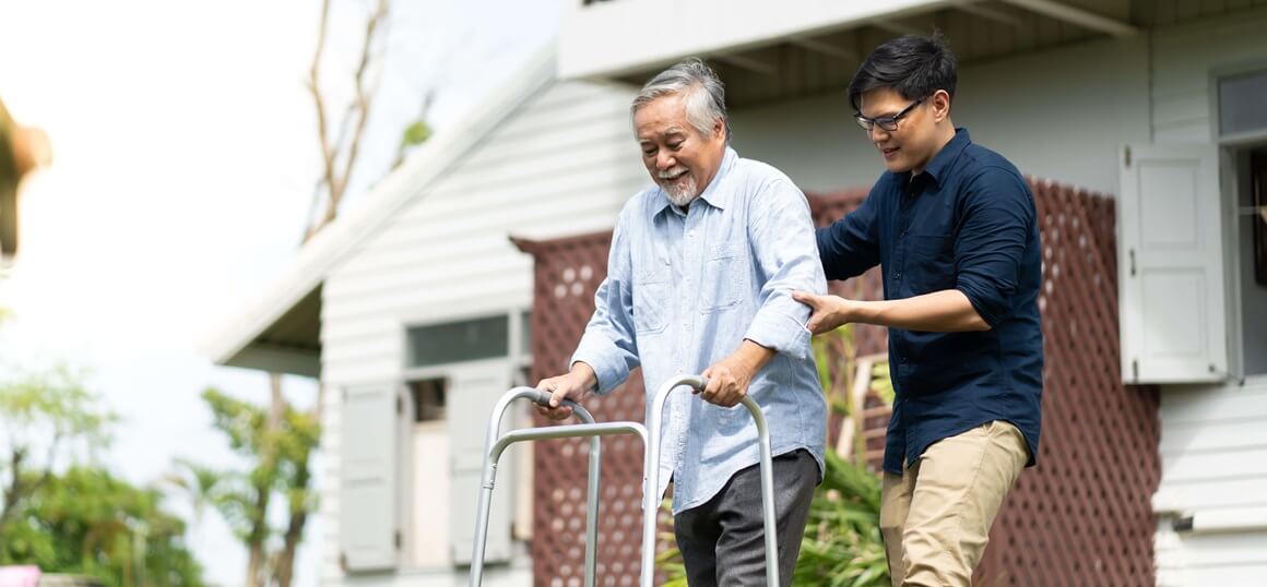 An older adult using a walker is assisted by a younger caregiver.