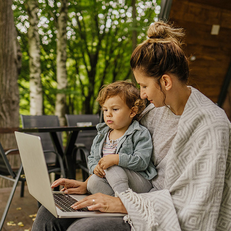 Woman uses her laptop to enroll in health coverage through Maine’s state-based marketplace. She is sitting outdoors on a deck, with a blanket around her shoulders and a toddler in her lap.