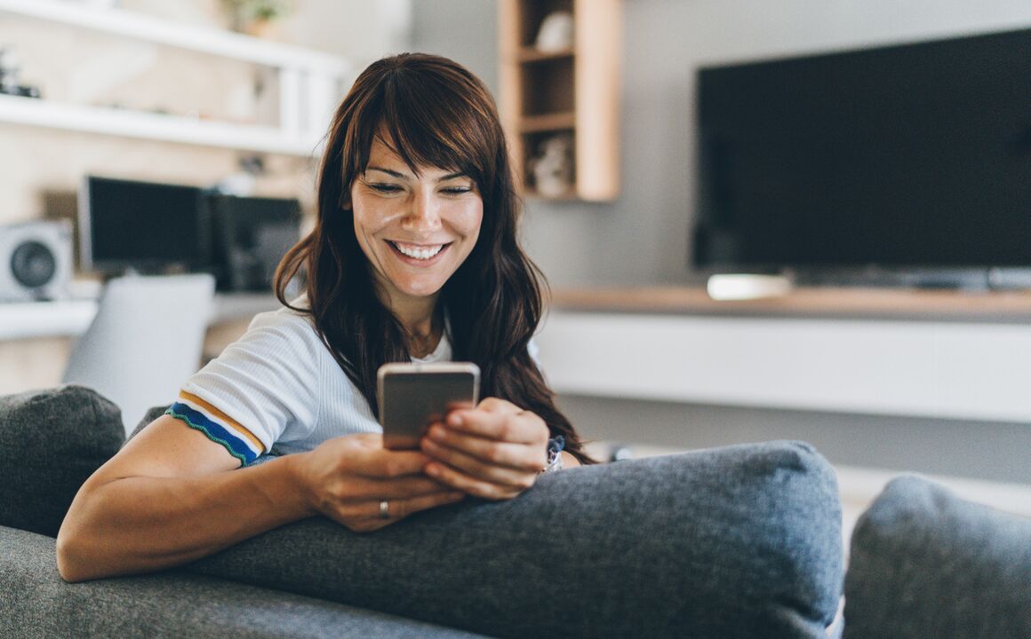 Woman leaning over the back of a sofa at home, using the smartphone in her hands to enroll in health insurance