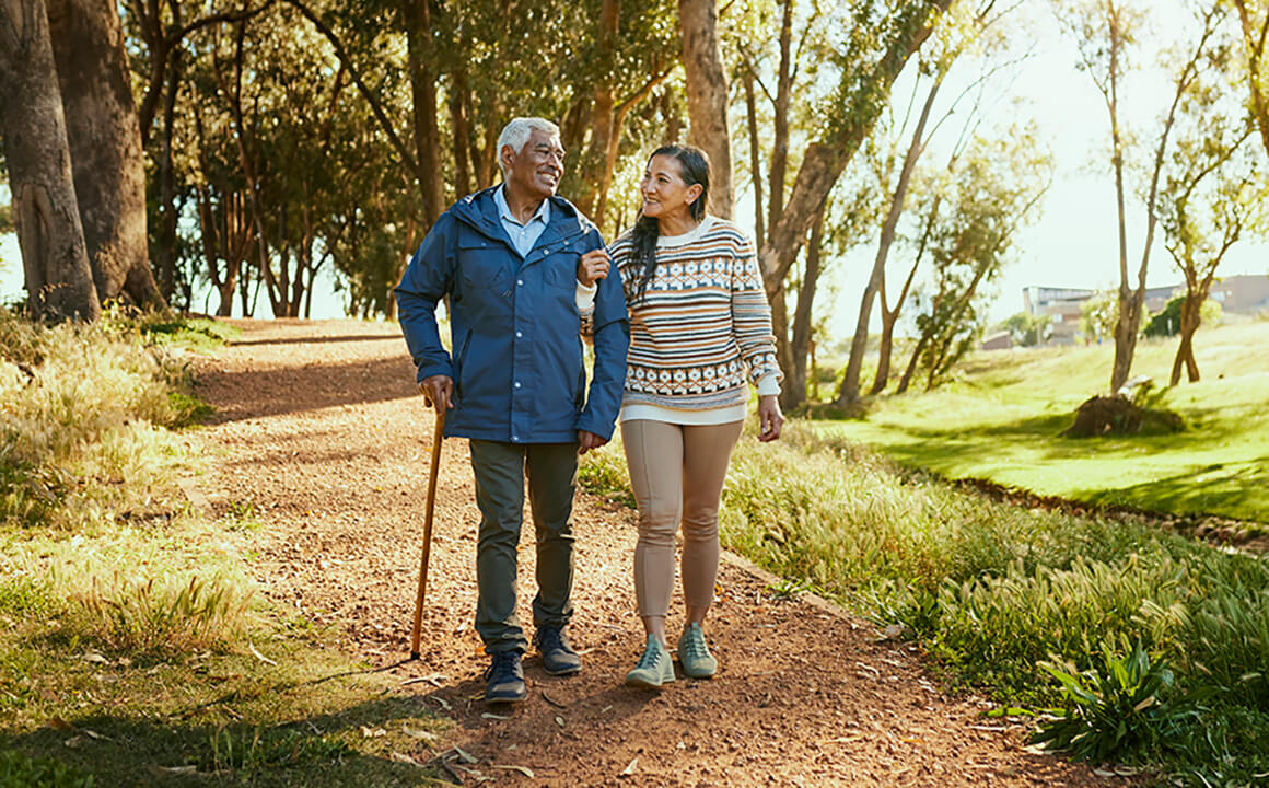 A family caregiver enjoys the outdoors with her father. He uses a cane as they walk on a path. They are both smiling. 