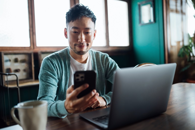 Man uses a smart phone to enroll in health coverage while sitting at a table with a laptop and coffee mug.